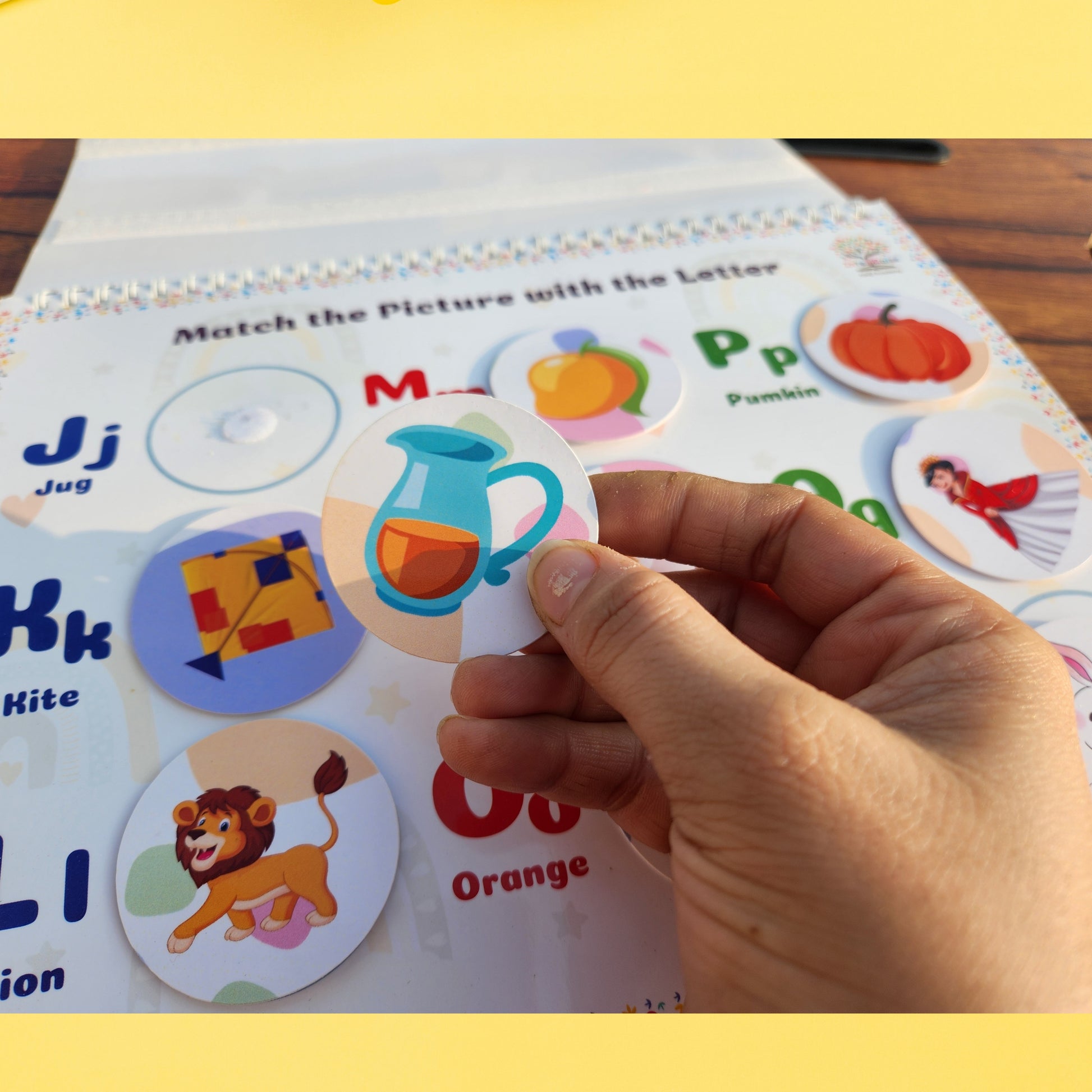 Hand holding a letter 'J' sticker over a matching game with colorful letters and pictures. sticker match book
